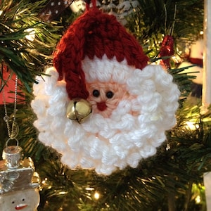 May include: A crocheted Santa Claus ornament with a red hat, white beard, and a gold bell. The ornament is hanging on a Christmas tree.