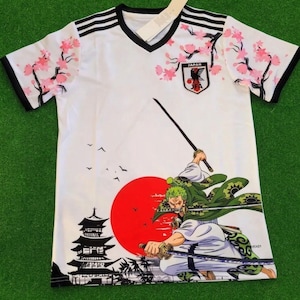 Peut inclure: Un t-shirt blanc à col en V avec des rayures noires sur les épaules, des branches de cerisiers roses et l'emblème de la Japan Football Association. Le motif montre un épéiste aux cheveux verts avec trois épées, un soleil rouge et une pagode.