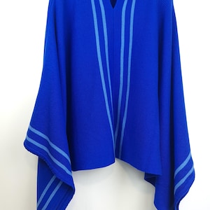 Könnte beinhalten: Ein leuchtend blaues Poncho-Oberteil mit Kragen und V-Ausschnitt. Das Oberteil hat hellblaue Streifen auf der Vorderseite und an den Ärmeln. Das Kleidungsstück wird auf einem schwarzen Kleiderbügel präsentiert.