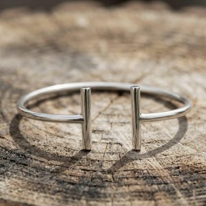 May include: A polished silver-toned cuff bracelet featuring a circular band that terminates in two vertical, perpendicular bars at the opening. The metal surface is smooth and reflective, showcasing a clean, open-ended design structure.