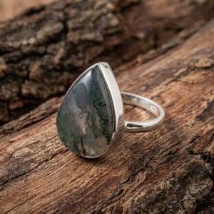 May include: A teardrop-shaped moss agate gemstone ring set in a polished silver band. The stone features translucent grey and green tones with dark, branch-like inclusions throughout. The metal band is stamped with 925 to indicate sterling silver.