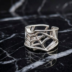 May include: A silver-toned ring featuring an openwork design that resembles a spider web. The band has a wide, structured appearance with interconnected geometric lines forming a lattice pattern throughout the entire circumference of the ring.