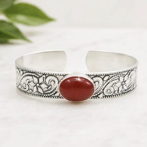 Puede incluir: Una pulsera tipo brazalete de tono plateado con diseño abierto y un patrón floral y de volutas en relieve. Una piedra lisa de forma ovalada y color rojo intenso está engastada en el centro, contrastando con el acabado metálico plateado.
