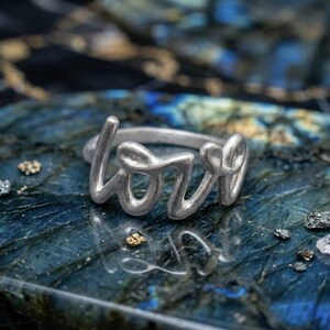 Puede incluir: Un anillo de metal en tono plateado que presenta la palabra love en una fuente cursiva continua. El anillo tiene un acabado mate cepillado y una banda redondeada que se conecta con las letras, creando una pieza de joyería cohesiva.