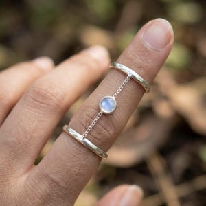 May include: A silver ring connected by a delicate chain to a smaller ring, with a round, light blue gemstone in the center. The ring is worn on a finger, showcasing its minimalist design and elegant style.