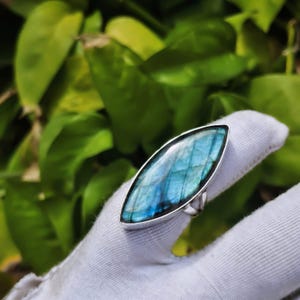 May include: A marquise-shaped labradorite gemstone ring set in a polished silver band. The stone displays a vibrant blue iridescence with visible internal fractures and a smooth, domed surface, securely held by a simple metal bezel setting.