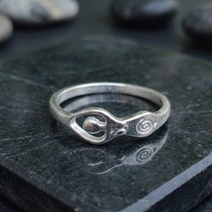 May include: A silver-toned ring featuring a stylized figure with a rounded head and a spiral design on the side. The band is smooth and circular, showcasing a metallic finish that reflects light across its surface, creating a distinct contrast.