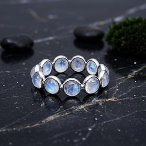 May include: A silver ring featuring a series of round, iridescent moonstone gemstones. The gemstones have a milky white base with blue flashes. The ring is set against a dark, reflective surface.