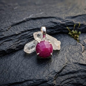 Könnte beinhalten: Ein runder, facettierter, tief pink-roter Edelsteinanhänger in einer silberfarbenen Metallfassung mit Krappen. Der Anhänger ist vor zwei klaren, länglichen, spitzen Quarzkristallen platziert und weist eine polierte, reflektierende Oberfläche auf.