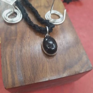 May include: A black oval gemstone pendant set in a silver frame on a black braided cord necklace, accompanied by two silver spiral-shaped earrings with flat, textured tops. The jewelry set is displayed on a rectangular wooden block.