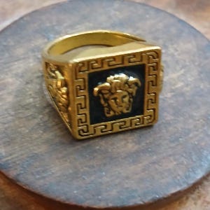 May include: A gold-colored ring with a square face featuring a black center and a Medusa head design. The ring has a Greek key pattern around the black square and on the sides. The ring is on a wooden surface.