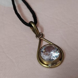 May include: A pendant necklace featuring a large, clear, round gemstone set in a gold-toned metal teardrop frame. The pendant hangs from a black cord, creating a simple yet elegant design. The gemstone sparkles, catching the light.