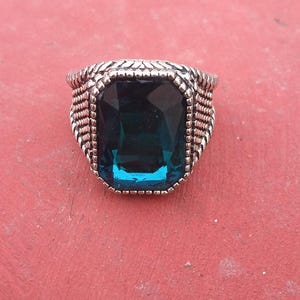 May include: A silver ring with a large, rectangular, teal-colored gemstone. The ring has a textured band with a woven design around the gemstone. The ring is set against a red background.
