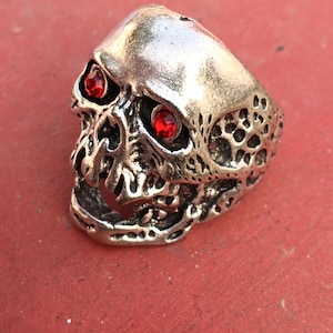 May include: A silver-colored skull ring with red gemstone eyes. The ring has a detailed, textured design, with the skull's features clearly defined. The ring is set against a red background.