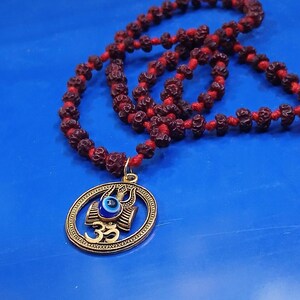 May include: A necklace featuring a string of dark brown, textured rudraksha beads on a red cord. A circular gold-toned pendant hangs from the cord, displaying a central blue and white evil eye bead, a trident symbol, and the Om symbol in gold.