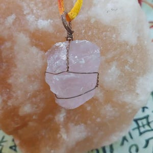 Może przedstawiać: Surowy, bladoróżowy wisiorek z kwarcem różowym, owinięty miedzianym drutem, zawieszony na sznurku z czerwonymi, pomarańczowymi i żółtymi nićmi. Wisiorek jest umieszczony na jasnopomarańczowym tle.