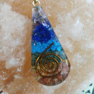 Może przedstawiać: Wisiorek w kształcie łzy z warstwami kolorowych kamieni i miedzianą spiralą. Wisiorek ma niebieskie, turkusowe, zielone i brązowe kamienie, zamknięte w przezroczystym tworzywie. Wisi na złotym łańcuszku.