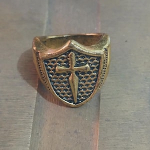 Puede incluir: Un anillo dorado con un diseño de escudo. El escudo presenta una cruz y un patrón texturizado. El anillo probablemente está hecho de metal y tiene una estética vintage o antigua.