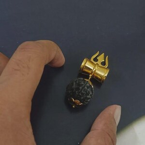 May include: A gold-toned metal pendant featuring a trident-shaped top attached to a cylindrical bail. A dark, spherical, textured bead is suspended from the bottom of the metal component, secured by a small gold-toned floral-shaped cap at the base.