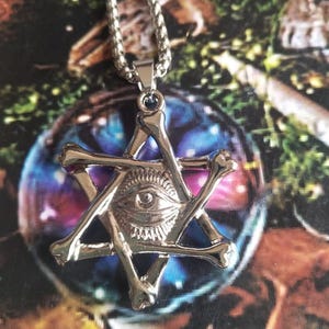 May include: Silver-toned pendant necklace featuring a six-pointed star design with bone-shaped elements. The center of the star has an eye symbol. The necklace has a chain.