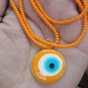 Peut inclure: Collier de perles orange avec un pendentif en forme d'œil. Le pendentif a un bord doré, un fond orange, un cercle intérieur blanc, un iris bleu et une pupille noire. Un accessoire de mode lumineux et coloré.