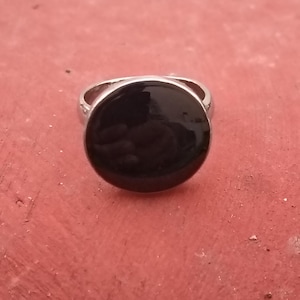 May include: A silver ring with a large, round, black stone. The ring band is a simple, polished silver. The black stone has a glossy finish, reflecting light. The ring is set against a reddish-orange background.