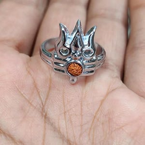 May include: A silver-colored ring featuring a trident design with a small, round, orange-brown stone at the center. The ring has a detailed, ornate design and is likely made of metal.