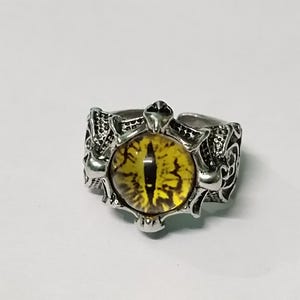 May include: Silver-toned ring featuring a large, yellow eye-shaped stone with black detailing. The ring has an ornate, textured design around the eye, with intricate patterns on the band. The ring is set against a white background.