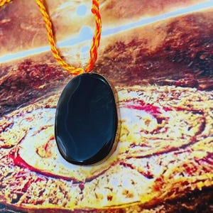 May include: A black oval pendant with a gold-colored border hangs from a yellow and red braided cord. The pendant is smooth and reflective. The background has a warm, earthy color palette.