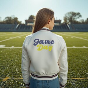 May include: A white satin bomber jacket with the words "Game Day" in blue and yellow script on the back. The jacket has a striped collar and cuffs. The person is standing on a football field.