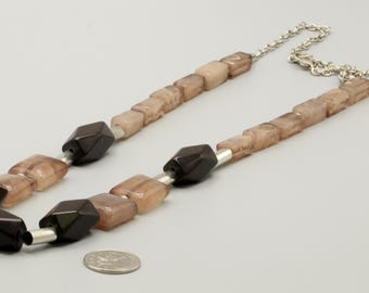 May include: A necklace featuring alternating dark brown faceted beads and translucent beige square beads, separated by silver-toned cylindrical spacers. The necklace includes a silver-toned chain and is shown next to a U.S. quarter for scale.