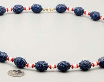 May include: A necklace featuring dark blue carved oval beads alternating with smaller white and red disc-shaped spacer beads. The strand is secured with a gold-tone metal spring ring clasp. A silver-colored coin is placed near the beads for scale.