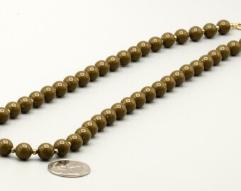 May include: A single-strand necklace composed of uniform, olive-green spherical beads separated by small gold-toned spacers. The necklace features a gold-toned circular clasp at the end and is displayed next to a silver-colored coin for scale.