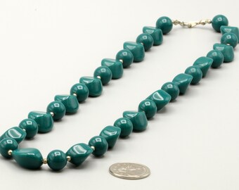 May include: A necklace composed of alternating teal-colored spherical and irregular geometric beads. The beads are strung together with small silver-toned spacers. A circular silver-colored coin is placed near the necklace for scale reference.