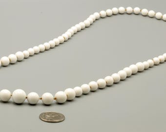 May include: A long strand of off-white spherical beads arranged in a graduated pattern, with larger beads centered and smaller beads toward the ends. A silver-colored US quarter coin is placed below the necklace for a size reference.
