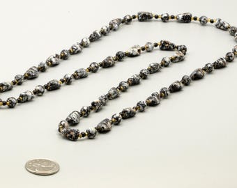 May include: A long necklace featuring irregularly shaped beads with a marbled black and white pattern, interspersed with small, round, metallic gold-colored spacer beads. A silver-colored coin is placed near the necklace for scale reference.