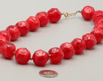 May include: A necklace composed of large, faceted, bright red beads with a glossy finish. The beads are strung together and secured with a gold-toned metal hook clasp. A small, round, silver-colored coin is placed near the necklace for scale.