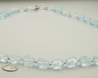 May include: A necklace composed of translucent, light blue, faceted, irregular-shaped beads separated by small, round, silver-colored metal spacers. A silver-colored United States quarter coin is placed next to the beads for scale reference.