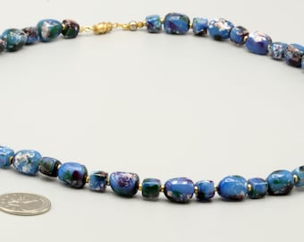 May include: A necklace composed of irregular, rounded blue glass beads with white and dark green marbled patterns. The beads are strung together with small gold-toned spacers and a gold-toned clasp. A silver-colored coin is placed near the necklace.