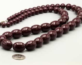 May include: A long strand of dark burgundy, oval-shaped beads with a smooth, glossy finish. The necklace is arranged in a circular loop next to a silver-colored coin for scale, showing the relative size of the beads in this single-strand piece.