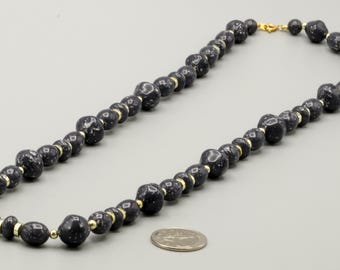 May include: A beaded necklace featuring dark blue, speckled, irregularly shaped beads interspersed with small, round, gold-colored metal spacers. The necklace is arranged in an oval shape next to a silver-colored coin for scale reference.