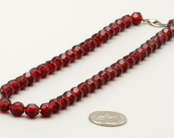 May include: A necklace composed of faceted, deep red translucent beads strung together, featuring a silver-toned metal lobster claw clasp. A standard United States quarter coin is placed nearby to provide a clear sense of scale for the beads.