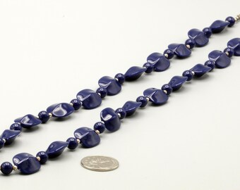 May include: A necklace featuring alternating dark blue, twisted, leaf-shaped beads and small, round, dark blue beads, accented with tiny metallic spacers. A silver-toned circular clasp secures the ends, and a US quarter coin provides scale.