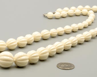 May include: A long necklace featuring off-white, spherical beads with a vertical ribbed texture. The beads are strung together in a single strand, and a silver-colored metal clasp is visible at the end. A US quarter coin is placed nearby for scale.