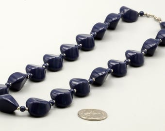 May include: A necklace featuring dark navy blue, irregularly shaped, faceted beads strung together with small silver-toned spacer beads. A silver-toned metal coin is placed near the necklace to provide a sense of scale for the bead size.