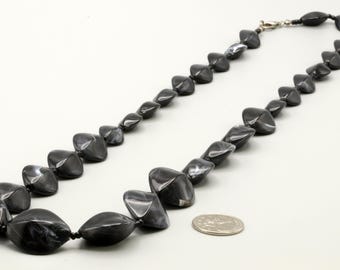 May include: A necklace composed of faceted, dark gray and black marbled beads with a glossy finish. The beads are strung together in a single strand, featuring a silver-toned metal lobster claw clasp. A US quarter coin is placed nearby for scale.