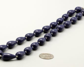May include: A necklace composed of smooth, dark blue, teardrop-shaped beads separated by small, round, silver-colored spacers. The necklace is arranged in a loop next to a silver-colored coin for scale, showing the size of the individual beads.