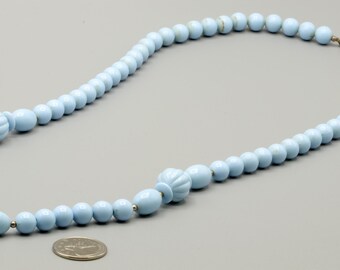 May include: A light blue beaded necklace featuring a series of smooth round beads interspersed with larger, ribbed, oval-shaped beads. The necklace is arranged in a circular shape next to a silver-colored coin for scale, showing the bead sizes.