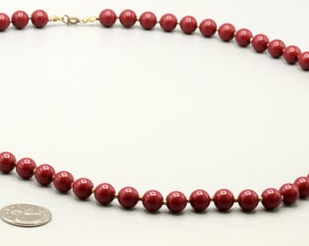 May include: A single strand necklace composed of uniform, smooth, deep red spherical beads. The necklace features a small gold-toned metal clasp at the top. A silver-colored coin is positioned near the bottom of the necklace for scale reference.