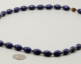 May include: A necklace featuring a series of smooth, dark blue, oval-shaped beads separated by small, thin, metallic gold-colored spacers. A silver-colored coin is positioned near the bottom of the necklace for scale reference.